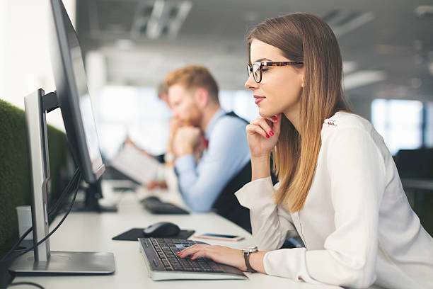 Female SEO specialist with glasses focused on desktop computer screen in modern open-plan office.