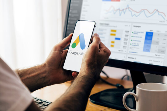 Person holding smartphone with Google Ads logo in front of a computer screen displaying Google Ads campaign dashboard.