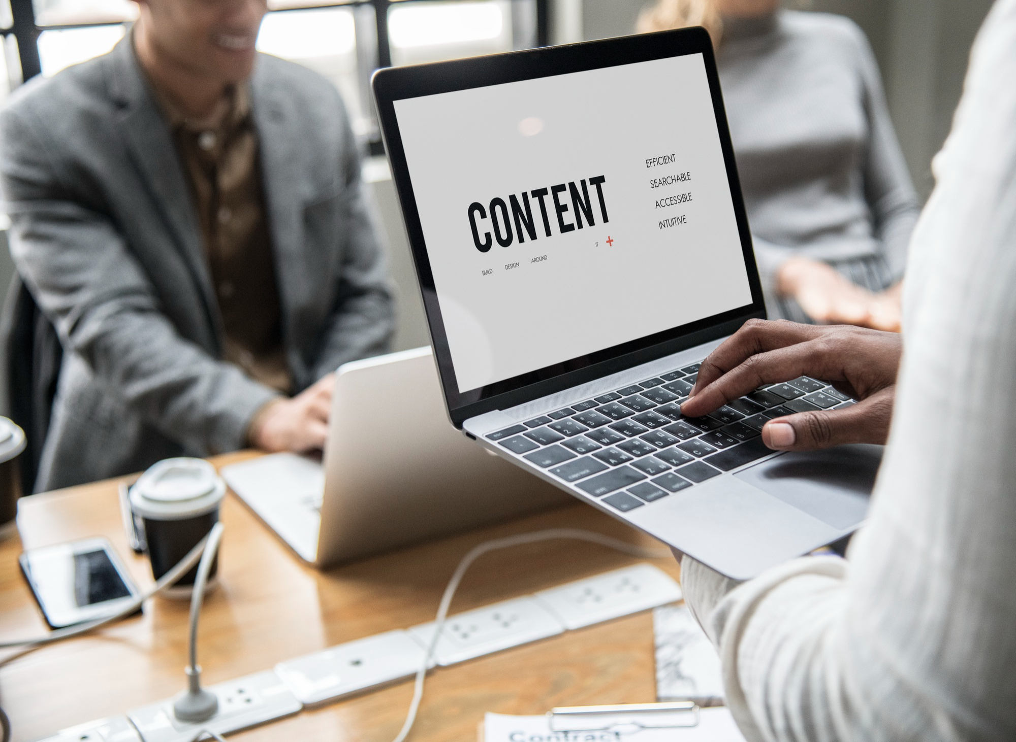 Person typing on a laptop headed CONTENT with words like searchable and accessible, colleagues nearby, developing a content strategy together.