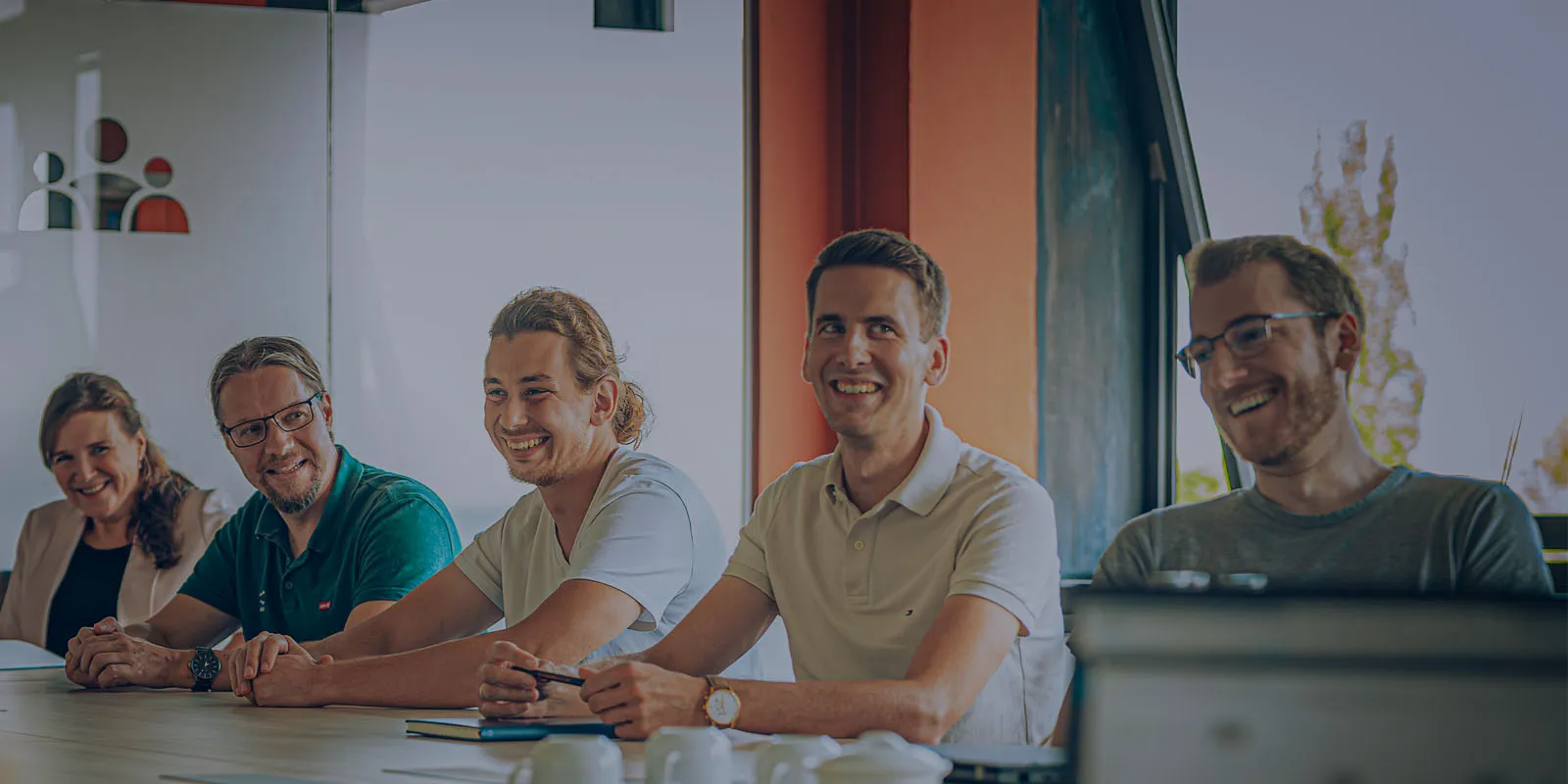 Fünf lächelnde Kollegen sitzen nebeneinander an einem Besprechungstisch in einem hellen Büro.