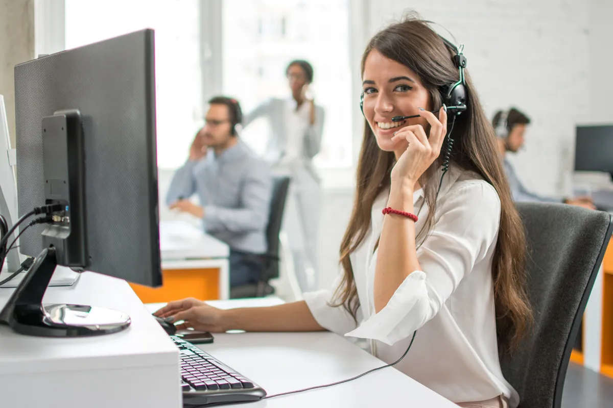 Junge Frau mit Headset lächelt im Büro vor Computer, im Hintergrund arbeiten Kollegen.