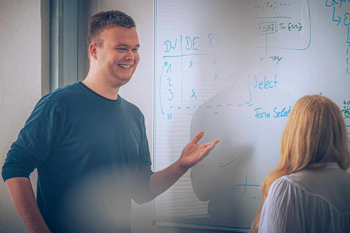Junger Mann erklärt einer Frau vor einem Whiteboard mit Diagrammen und Text etwas.