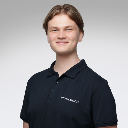 Junger Mann mit hellem Haar und schwarzem IP DYNAMICS Poloshirt lächelt vor grauem Hintergrund.