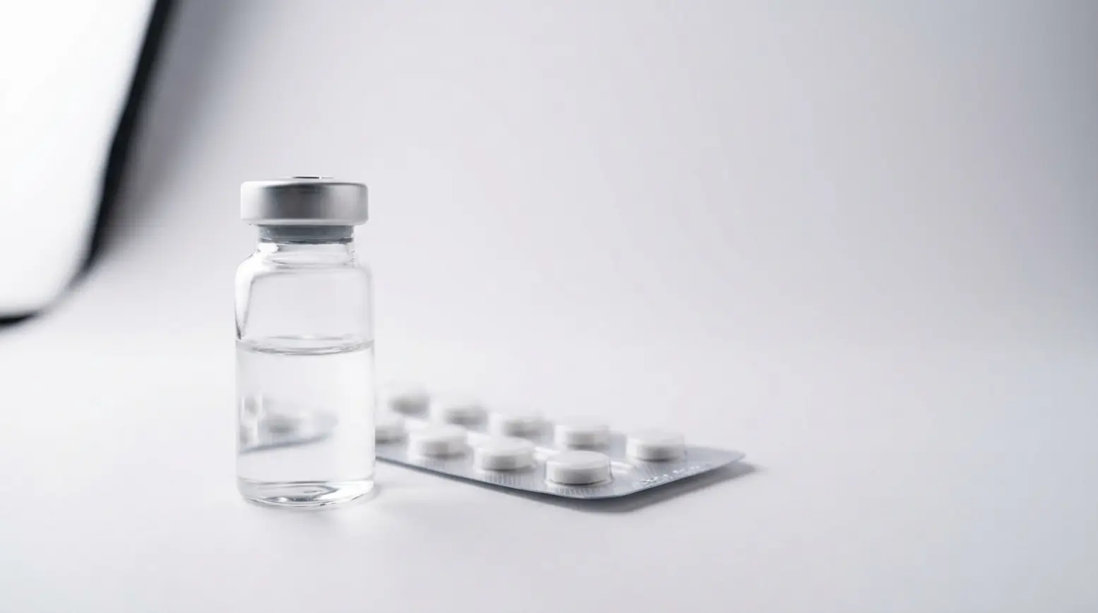 Clear glass vial with liquid and blister pack of tablets on a white surface, minimalistic style.