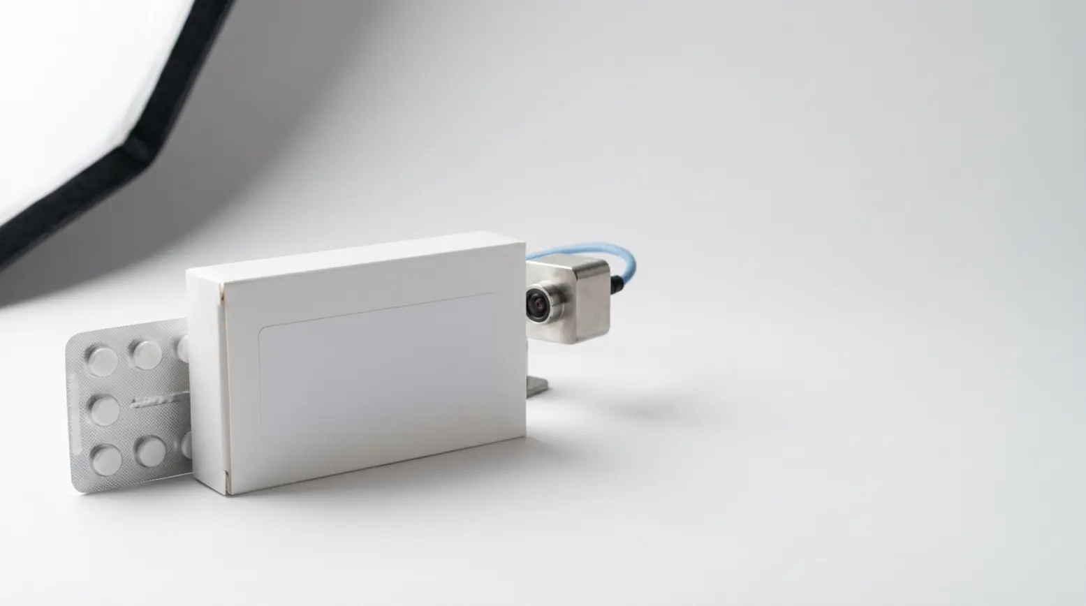 An unbranded white medicine carton and blister pack with a camera, set against a clean background.