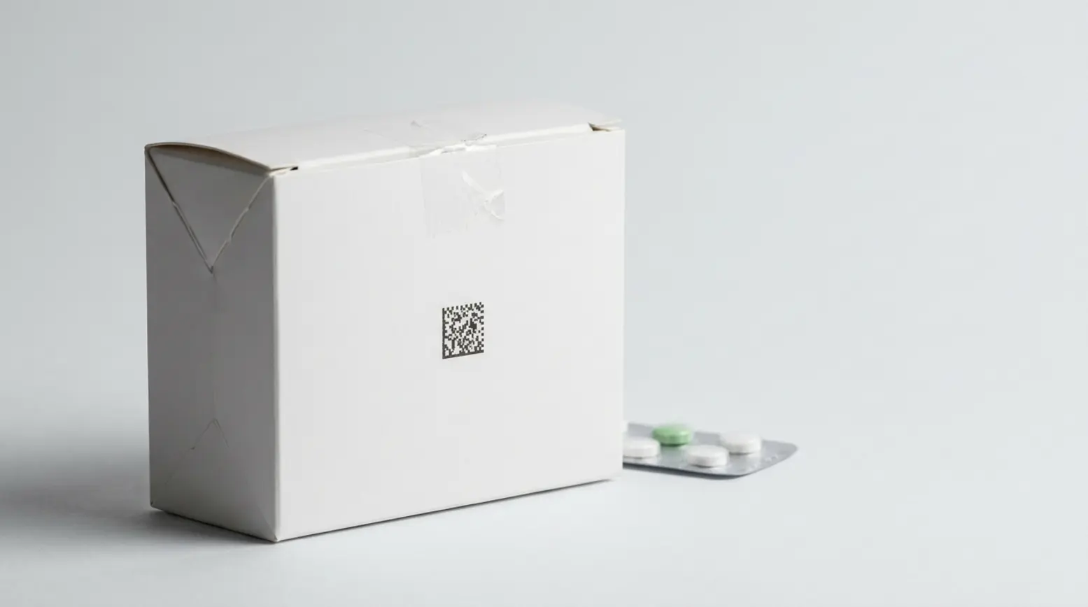 Clean white pharmaceutical box with tamper seal, blister pack, against soft grey background.