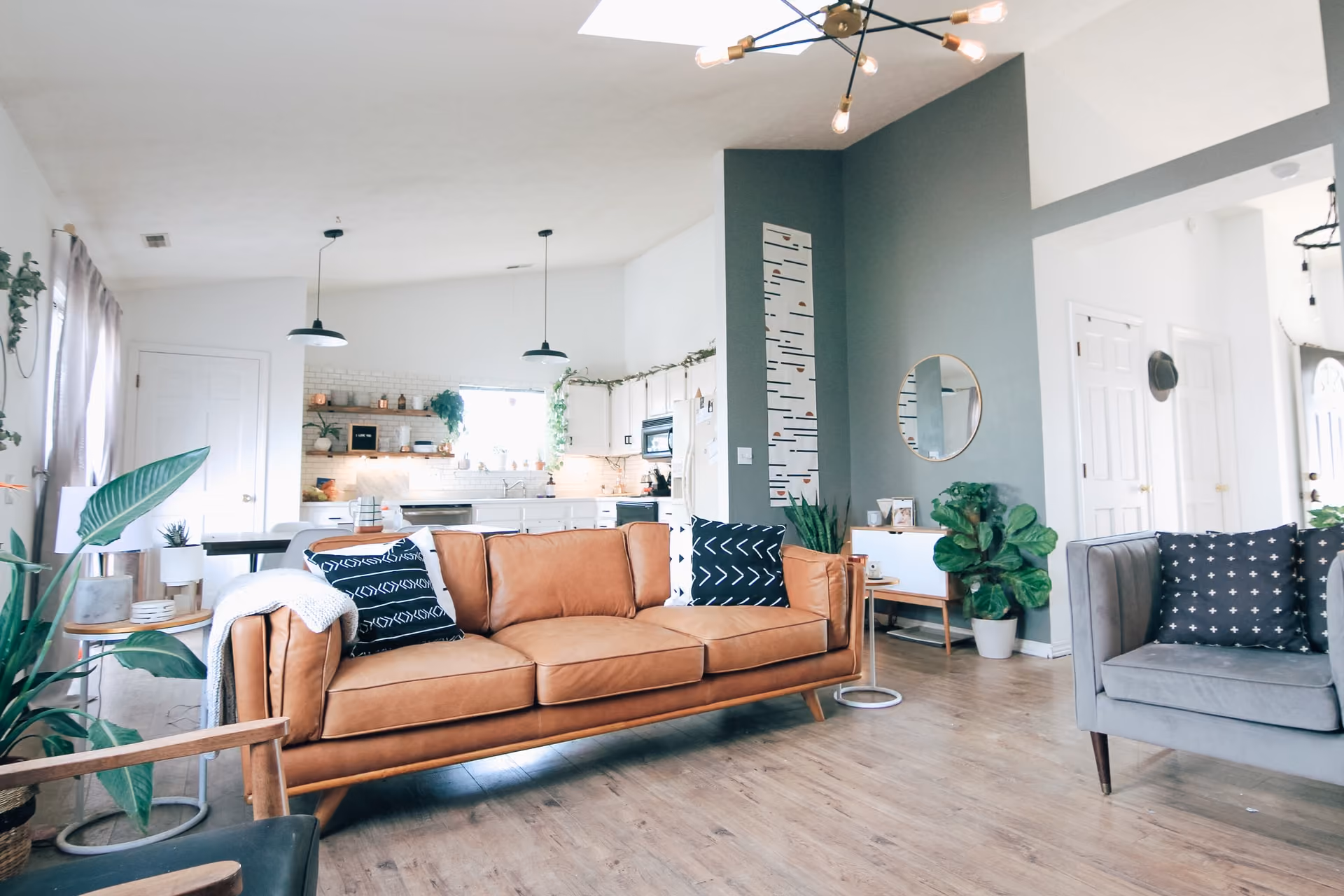Spacious modern living room with a tan leather sofa, gray armchair, wooden floor, and open kitchen in the background.