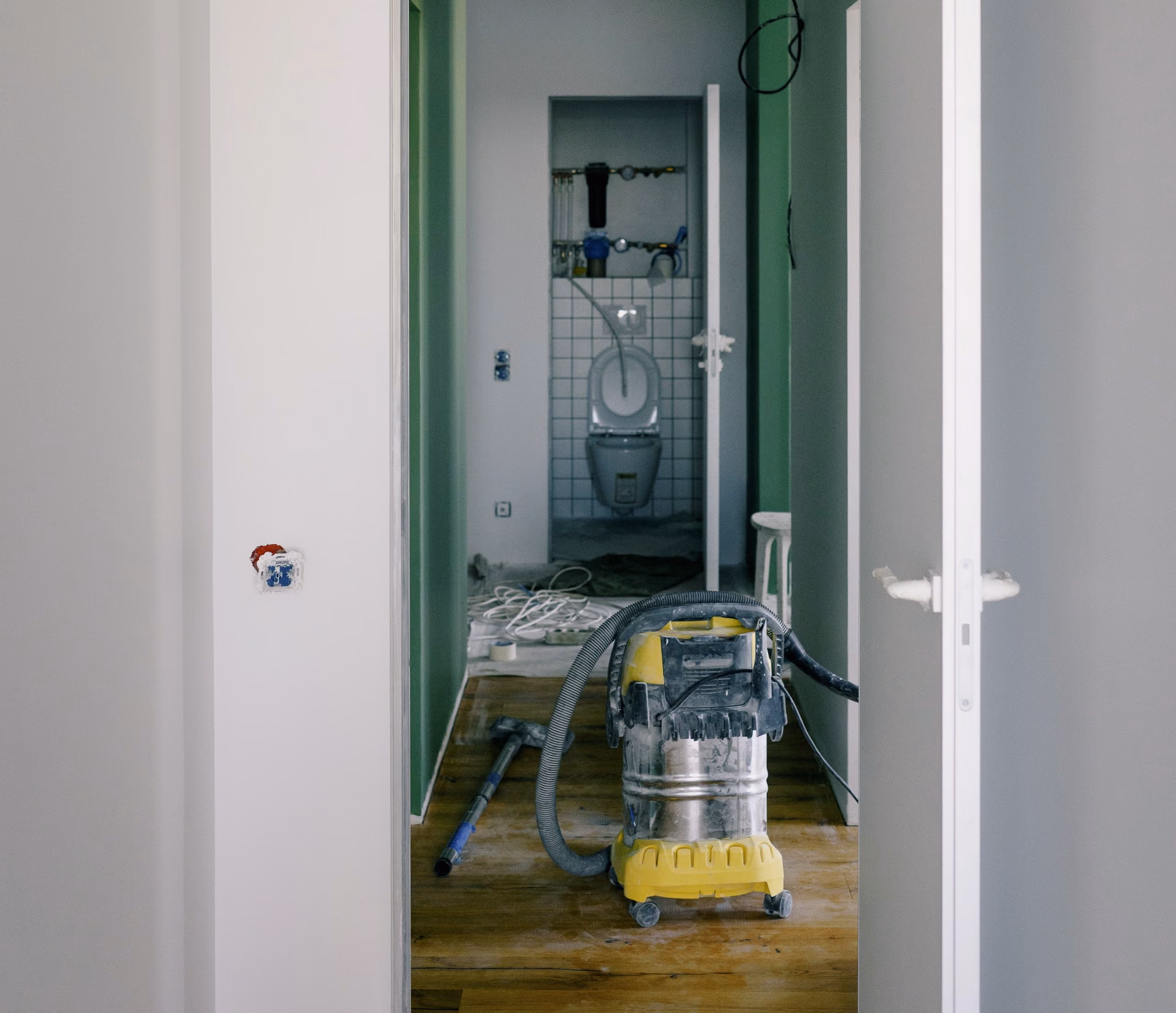 Vacuum cleaner and construction tools on wooden floor of an unfinished hallway leading to a restroom under construction.