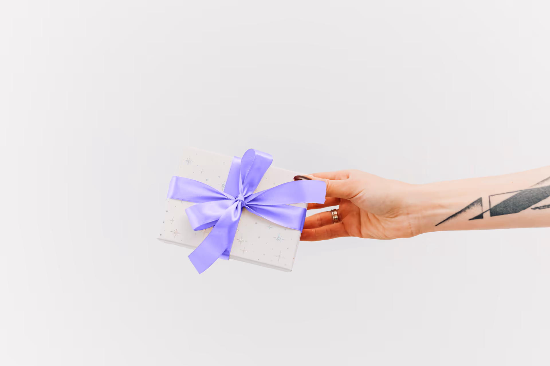 Mano extendida ofreciendo una caja de regalo blanca con moño morado, sobre fondo blanco.