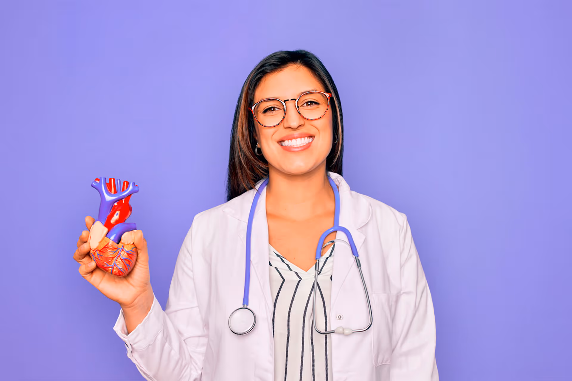 Doctora con bata blanca y estetoscopio mostrando una figura anatómica de un corazón humano, sobre fondo morado.