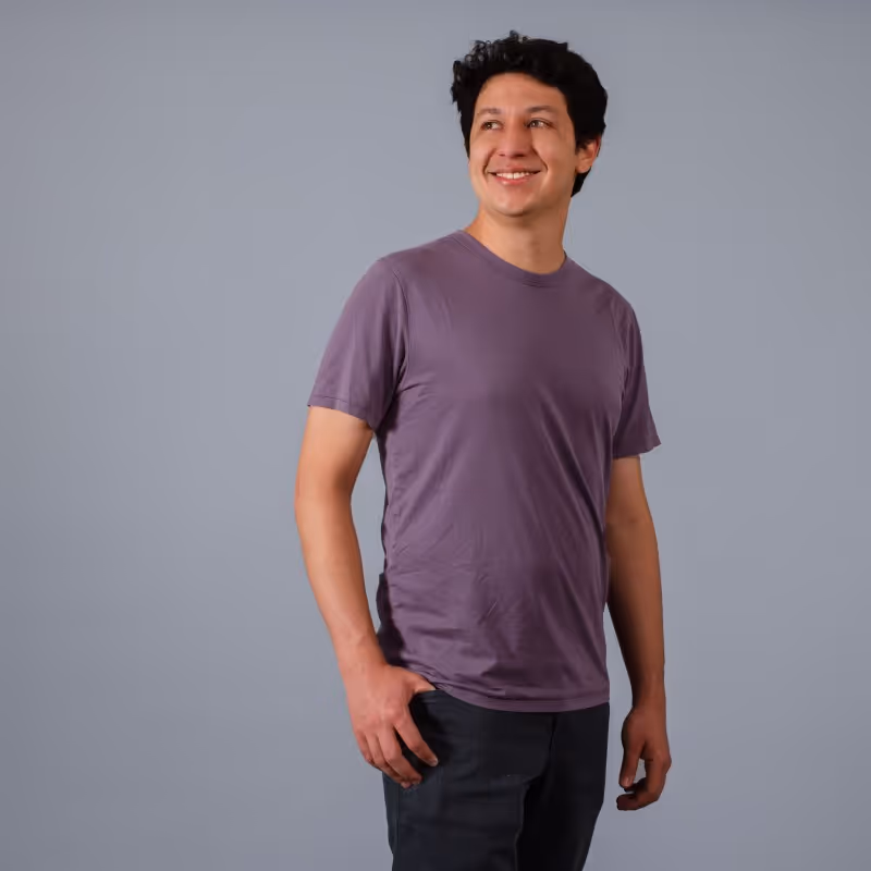Retrato de Sebastian Jimenez con camiseta morada y pantalón oscuro de pie sobre fondo gris, mirando hacia un lado y sonriendo.