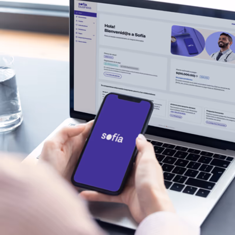 Persona sosteniendo un teléfono móvil con la app de Sofía abierta frente a una laptop que muestra el portal de Sofía Business, representando acceso digital a servicios médicos.