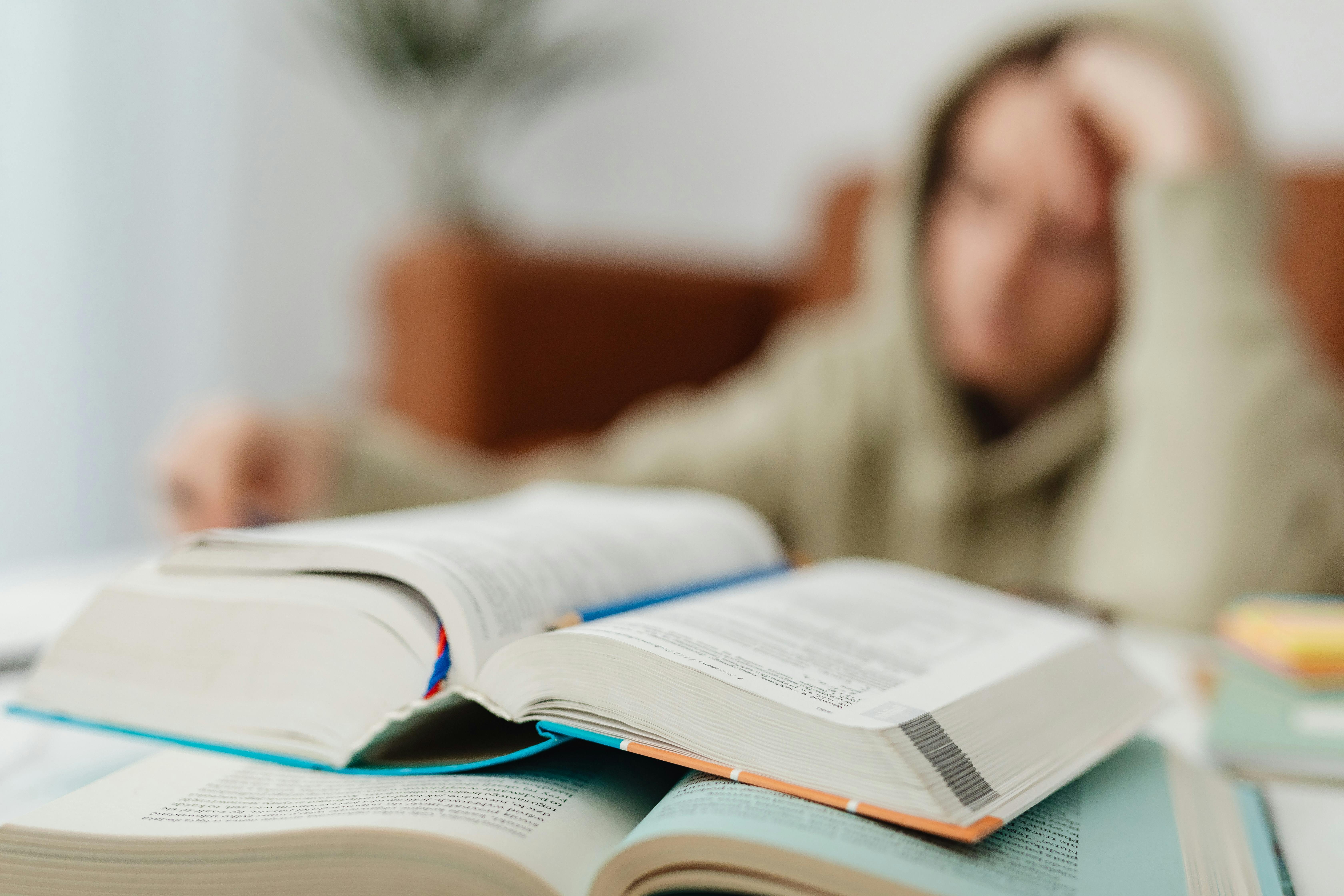 A student in a hoodie puts their hand to their forehead in frustration, with their books laid out on a table in front of them.