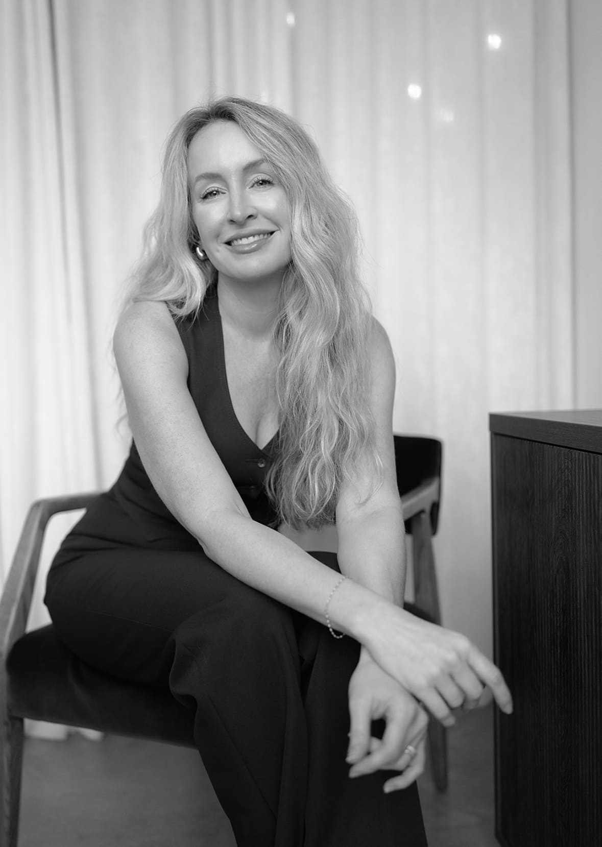 Smiling woman with long wavy hair seated on a chair wearing a sleeveless top and pants, with her hands resting on her knee.