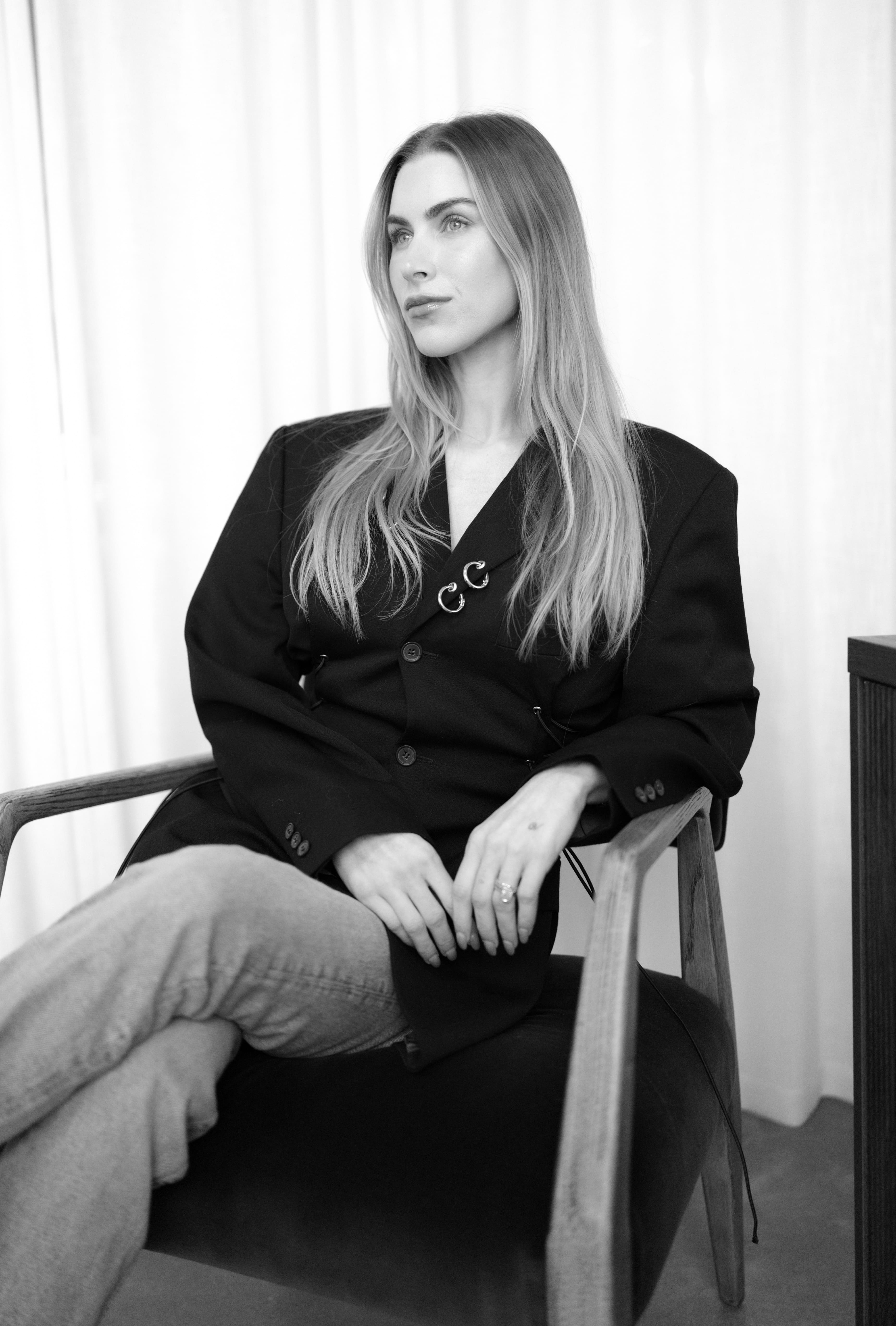 Black and white portrait of a woman with long hair sitting in a wooden chair wearing a black blazer and light jeans.