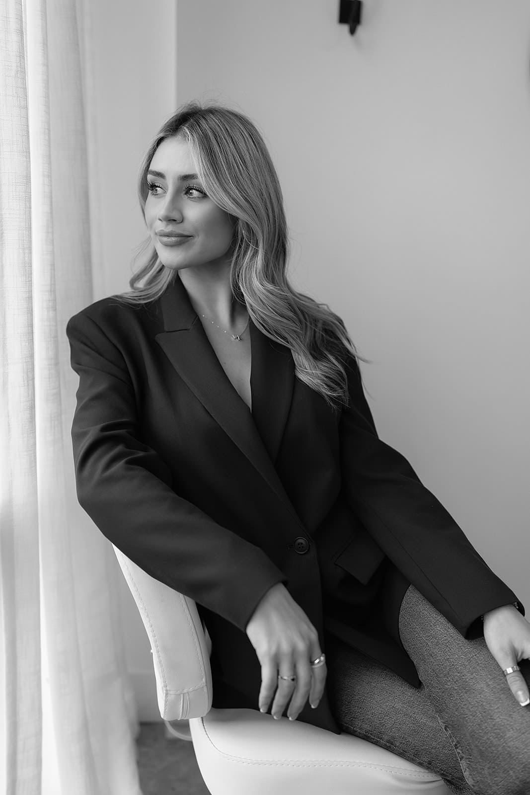 Young woman with long hair wearing a dark blazer and jeans, sitting on a white chair and looking out a window with curtains.