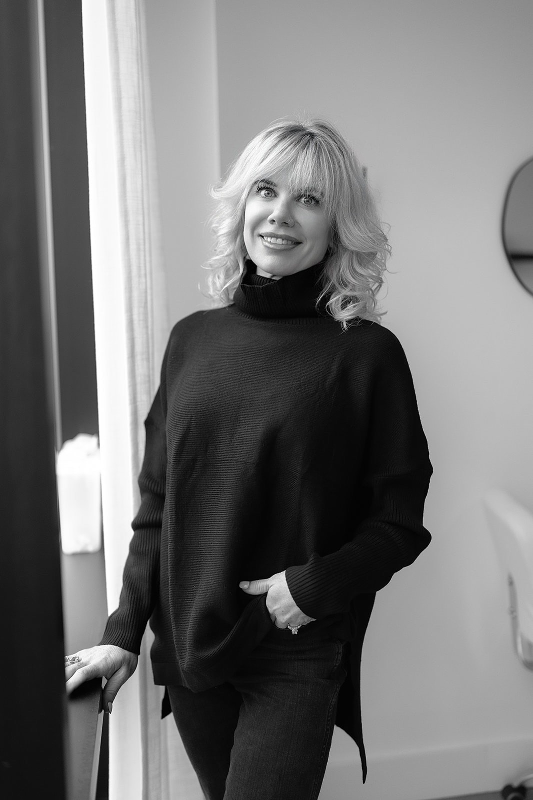Smiling woman with shoulder-length curly blonde hair wearing a black sweater, standing indoors near a window and looking slightly upward.