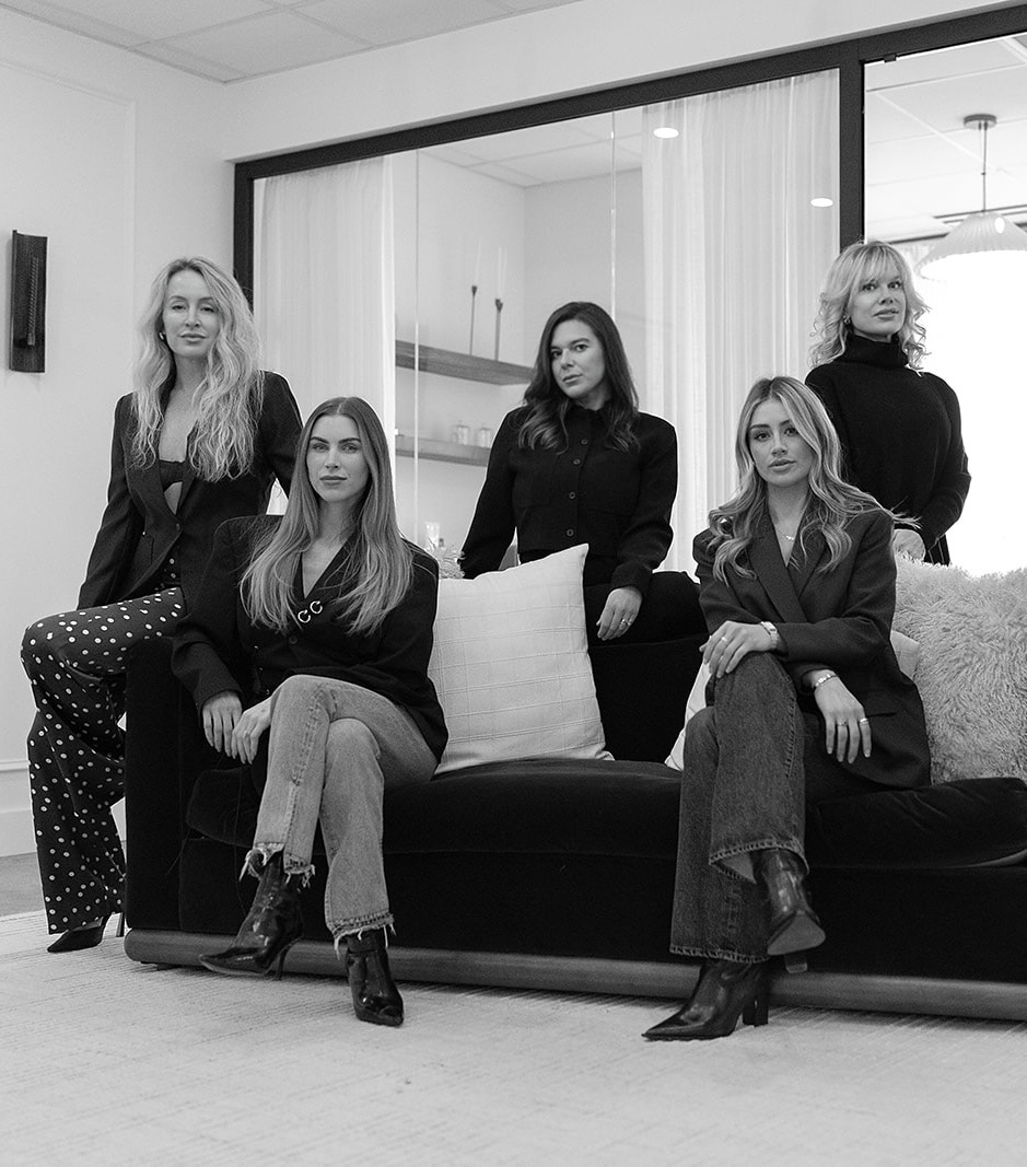 Five women posing in a modern indoor setting, three sitting on a black couch with pillows and two standing behind it, all dressed in stylish black and dark outfits.
