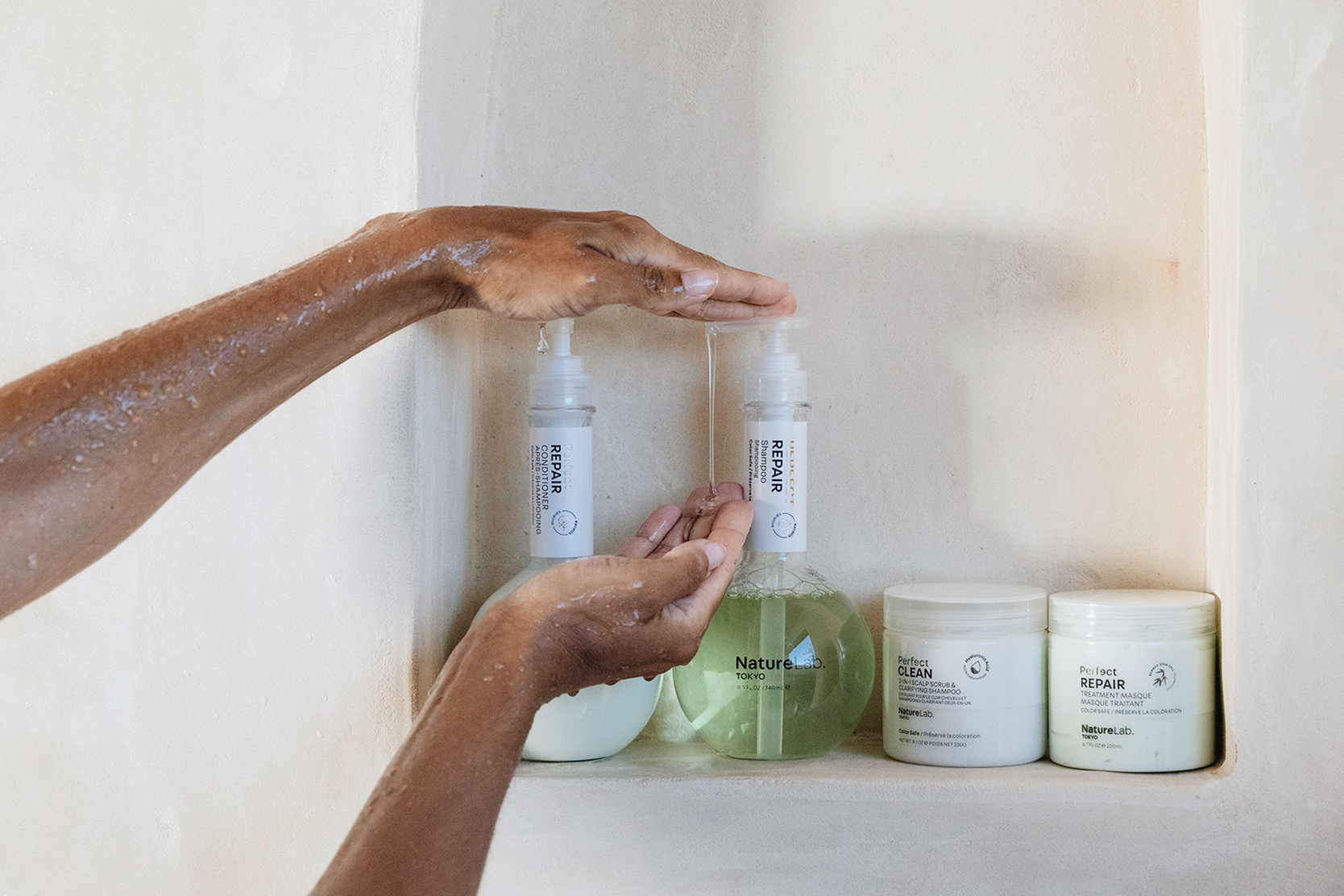 Wet hands dispensing liquid shampoo from a NatureLab Tokyo bottle in a shower niche with other hair care products.