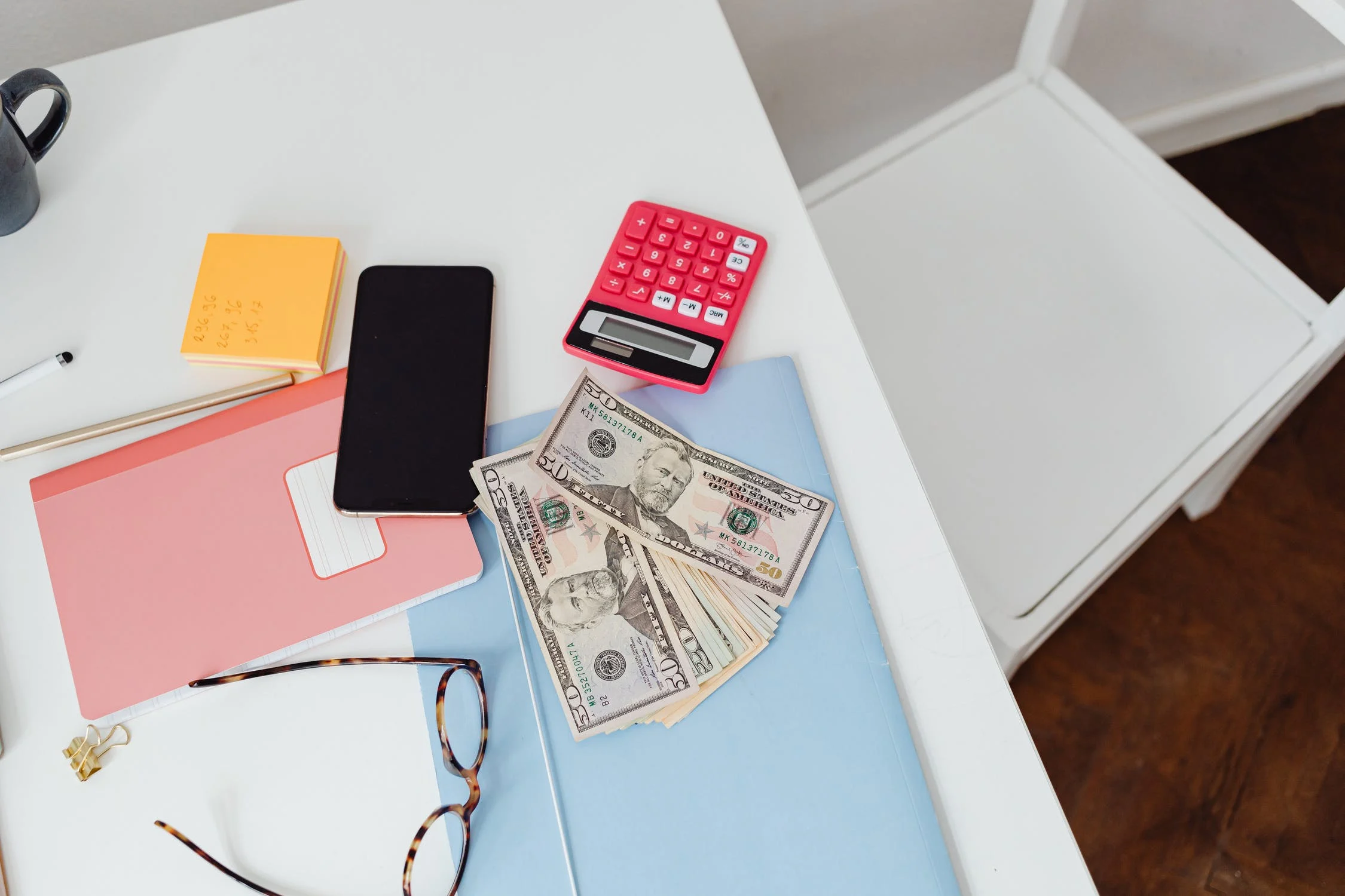 Miscellaneous items thrown askew on a white desk including: a notebook, a phone, post-it notes, a calculator, a couple of $50 bills, a folder, and some pens.