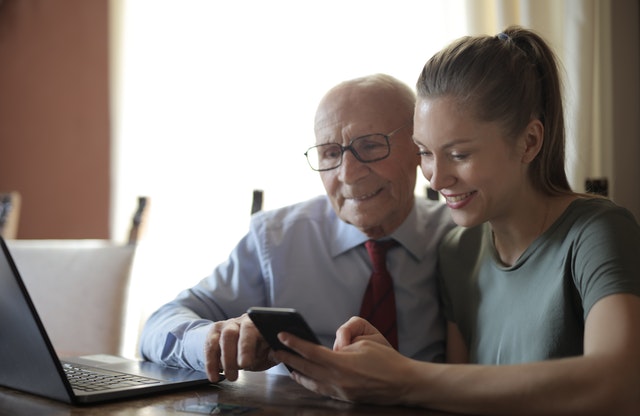 A daughter is seated next to her father at a table in front of a laptop. The daughter is holding a cell phone in her left hand, and the father is pointing at the phone screen.