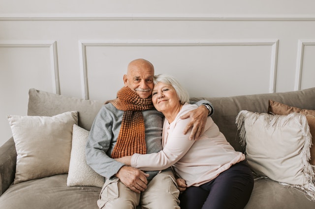 An older couple is seated on a couch. They have their arms around each other and are smiling and staring directly at the camera.