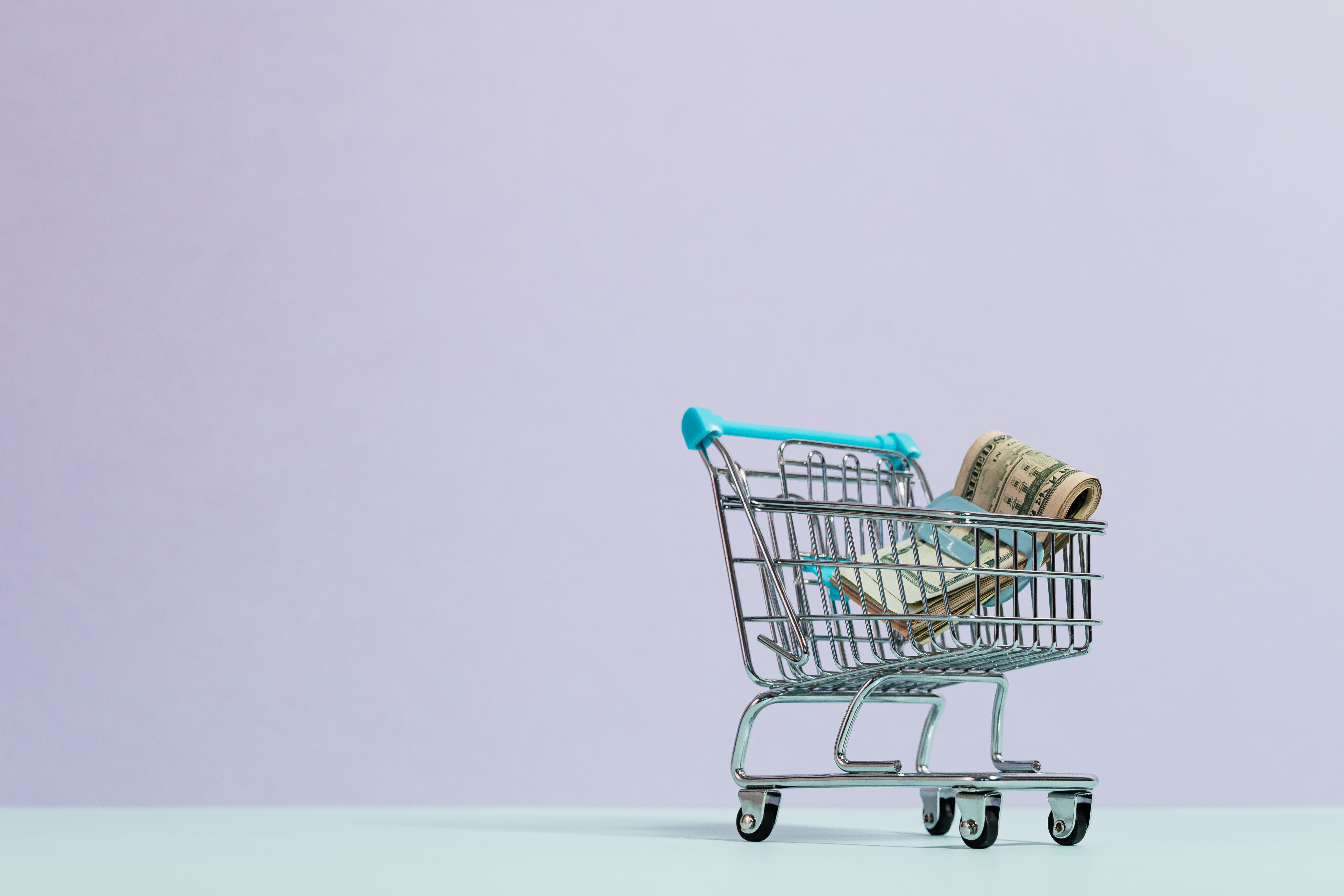 A miniature grocery cart that contains a wad of folded up dollar bills in front of a plain, lavender background.