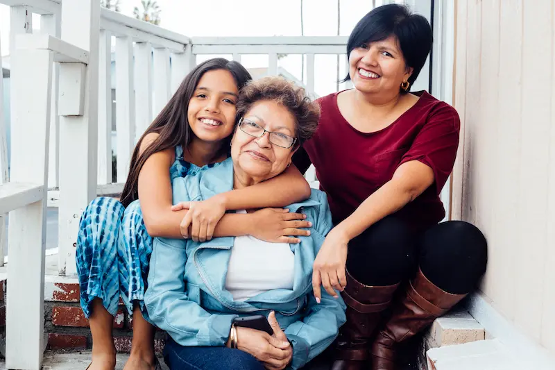 Smiling portrait of multi generational Latino family. Three women sit on stairs for a family photo, mom, daughter, and granddaughter.