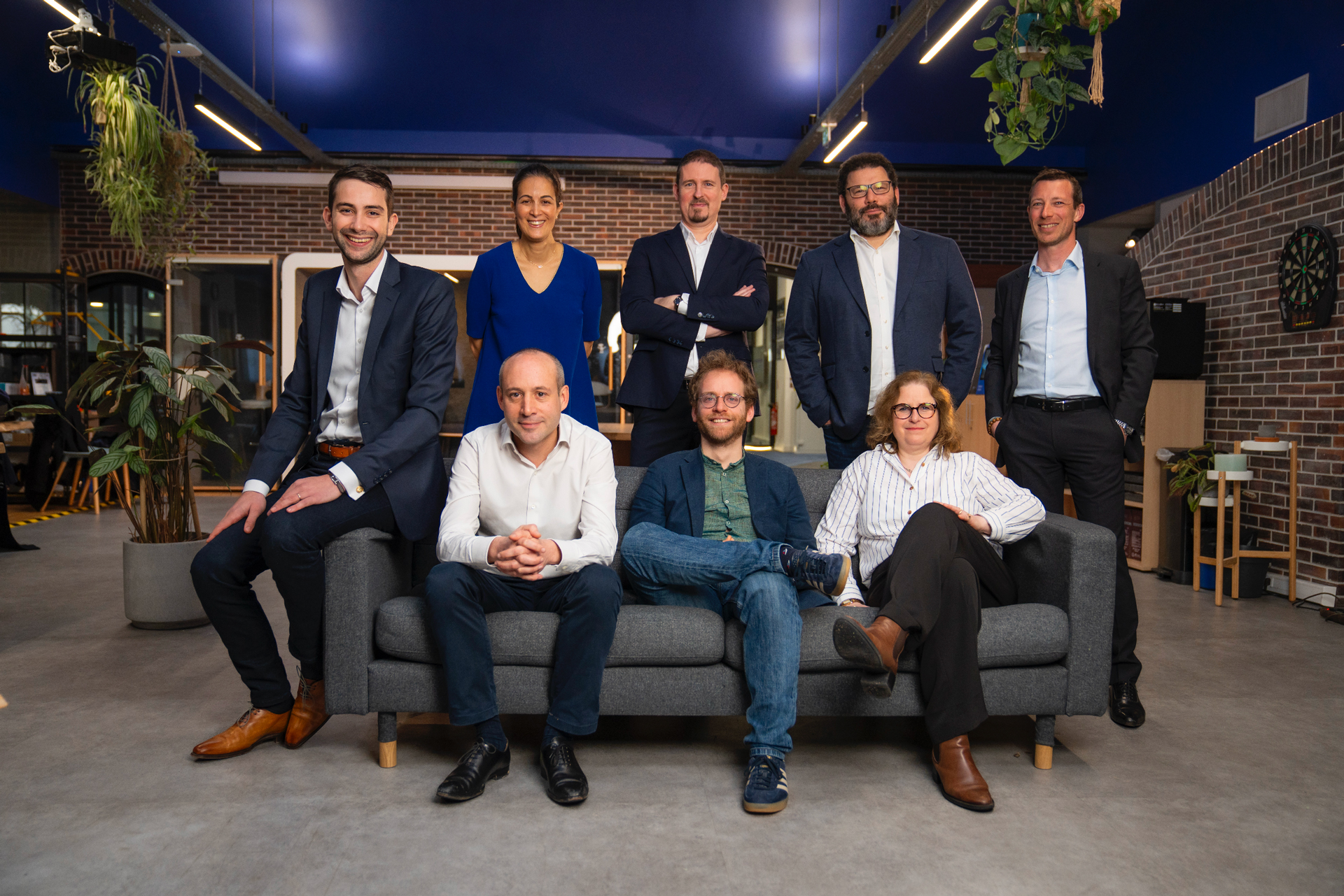 Group of nine professionally dressed people posing in an office with brick walls, some sitting on a gray sofa and others standing behind it.