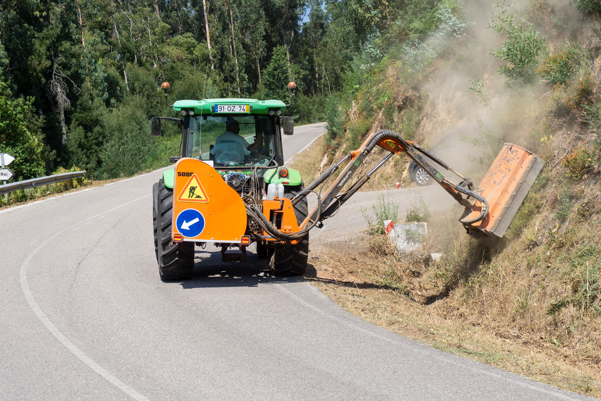 Trator verde com máquina azul e laranja cortando vegetação na berma de estrada curva rodeada por árvores.