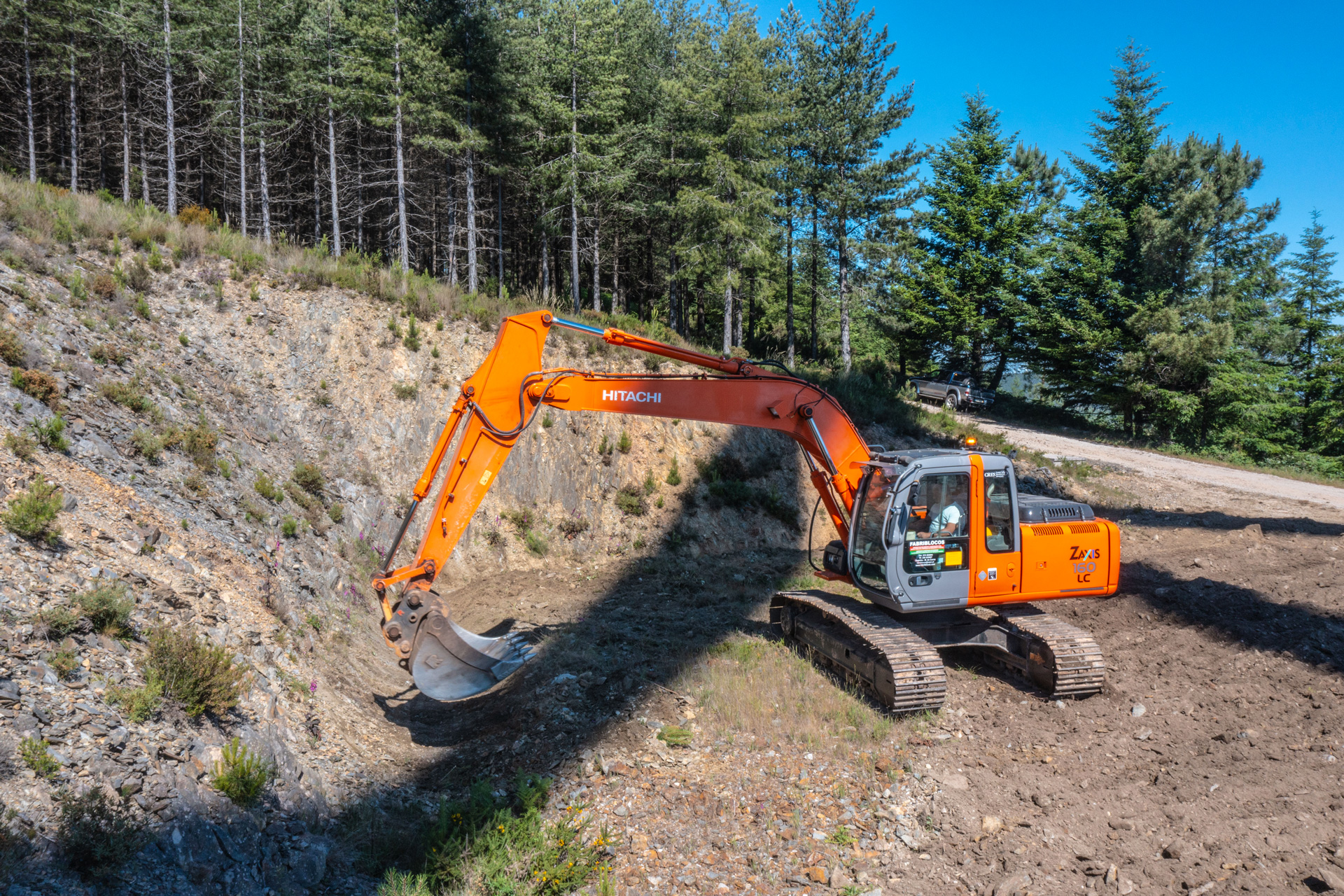 Escavadora laranja Hitachi a trabalhar num terreno rochoso com árvores de pinheiro ao fundo.