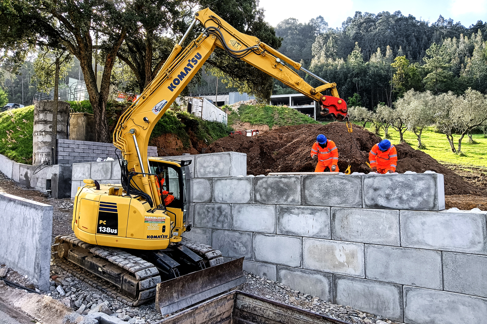 Escavadora amarela Komatsu a construir um muro de blocos de cimento com dois trabalhadores em equipamento de proteção laranja e capacetes azuis.
