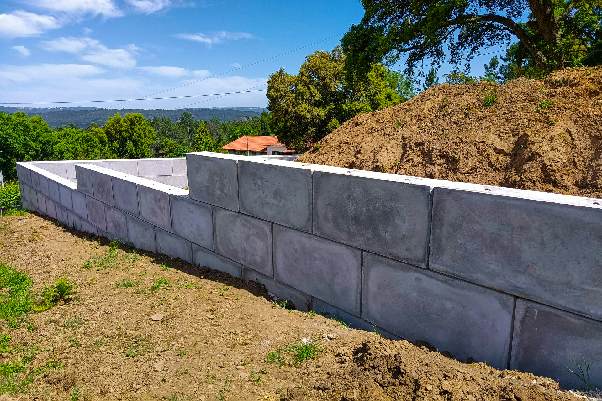 Muro de blocos de cimento parcialmente construído num terreno com terra e árvores ao fundo sob um céu azul.