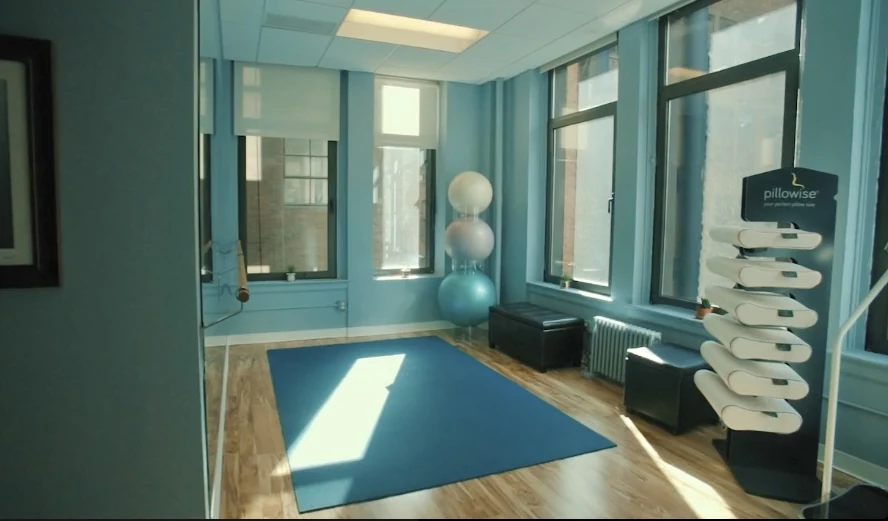 Sunlit movement studio with large windows, mirrored wall, exercise mat, and movement equipment