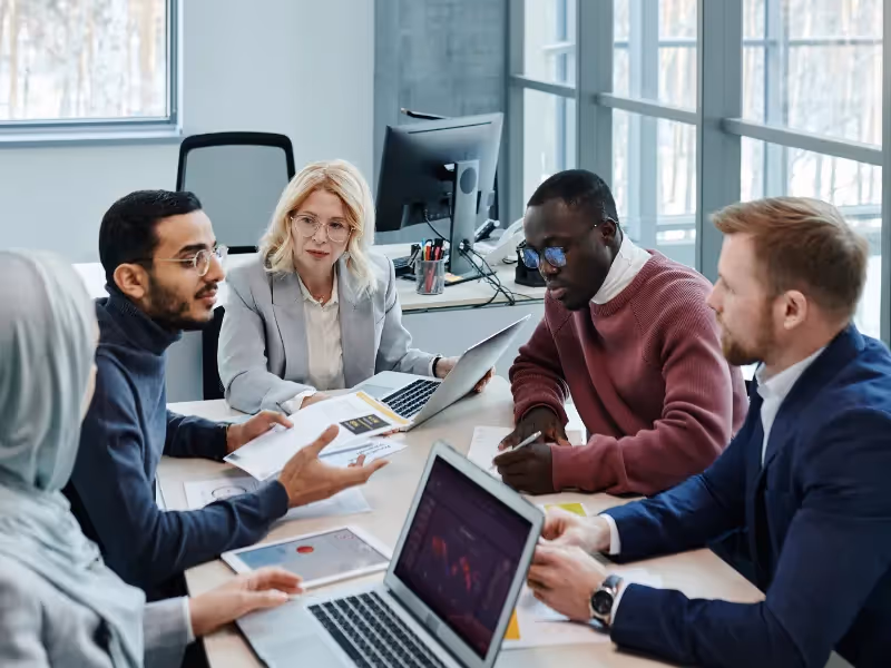 Larger image of an office room with employees having a discussion around a rectangular table