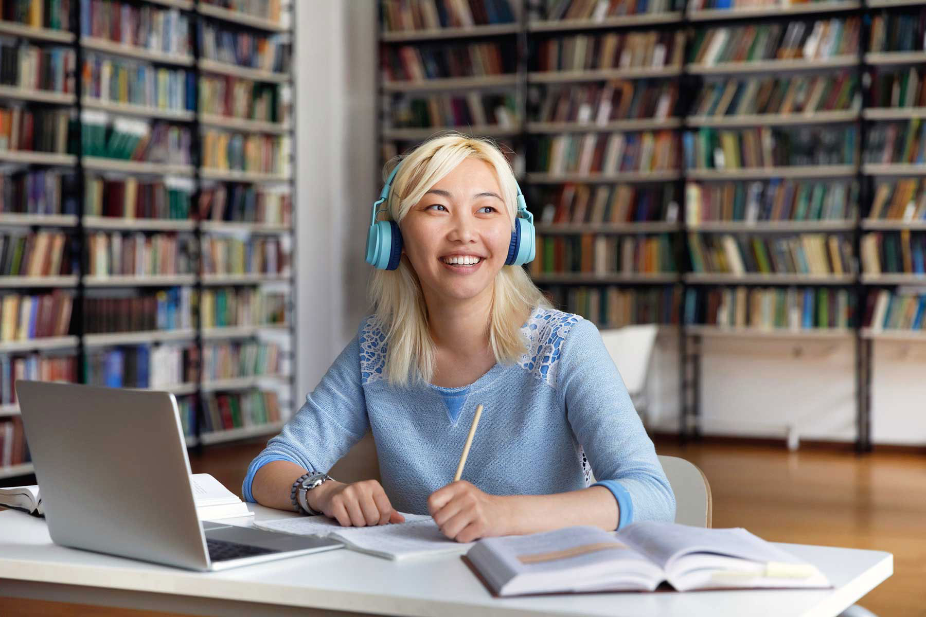 Smiling student wearing blue headphones studying with laptop and books in a library.