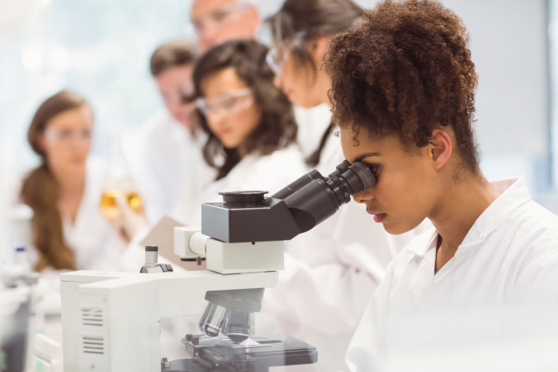 Young female scientist with curly hair looks through a microscope in a lab, with other scientists blurred in the background.