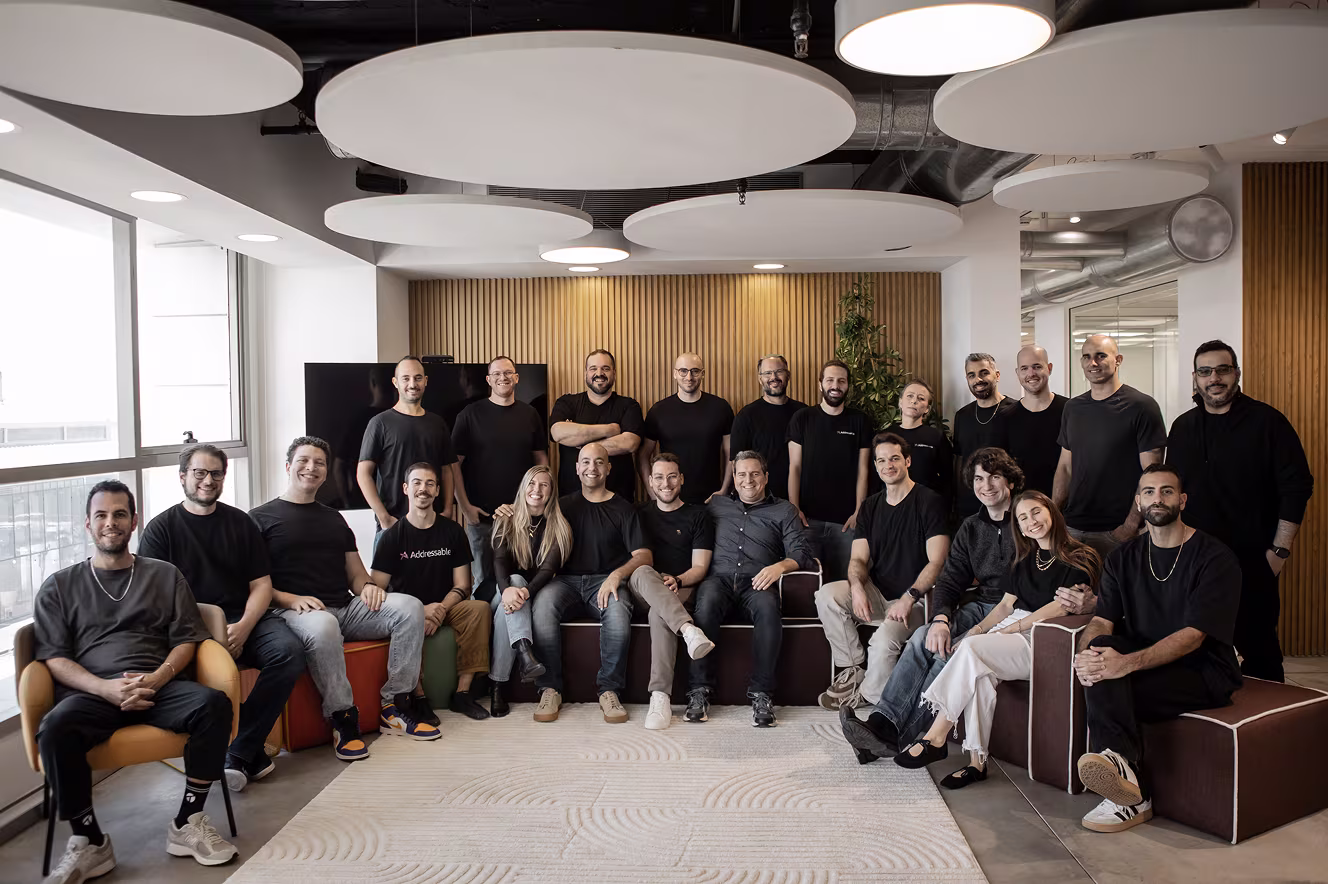 Group of 22 people posing and smiling in a modern office room with large windows and ceiling panels.