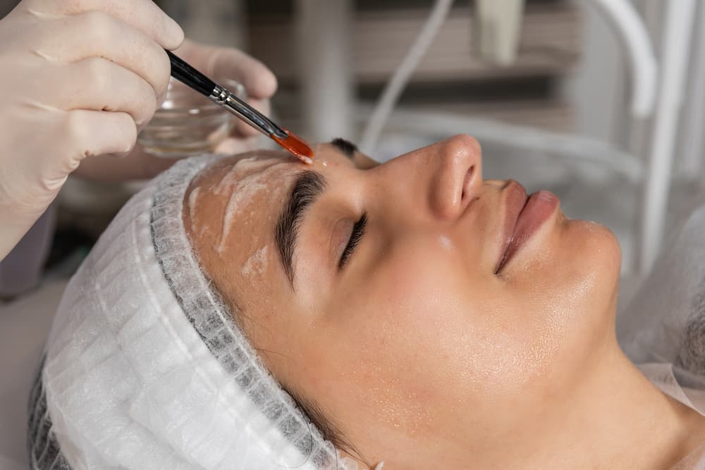 A professional applying a skin mask or peel to a woman's face during a regenerative dermatology anti-aging treatment.