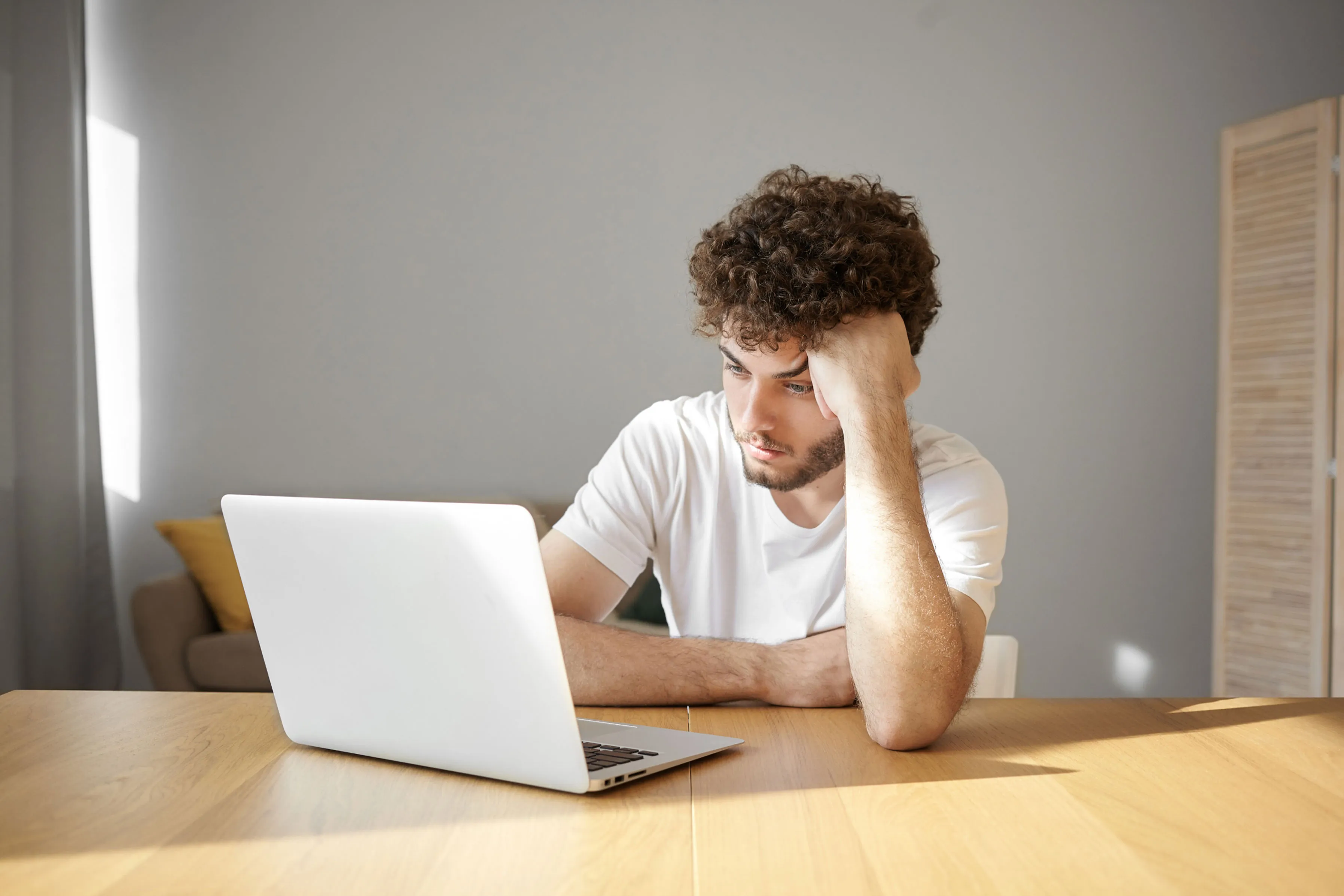Man with curly hair sitting at a table, looking at a laptop with a thoughtful expression.