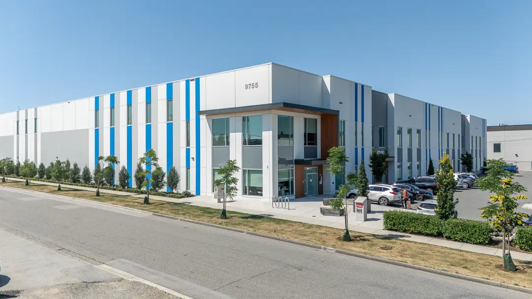 Modern industrial building with white and gray exterior accented by blue vertical stripes, a glass entrance, and parked cars in the lot.