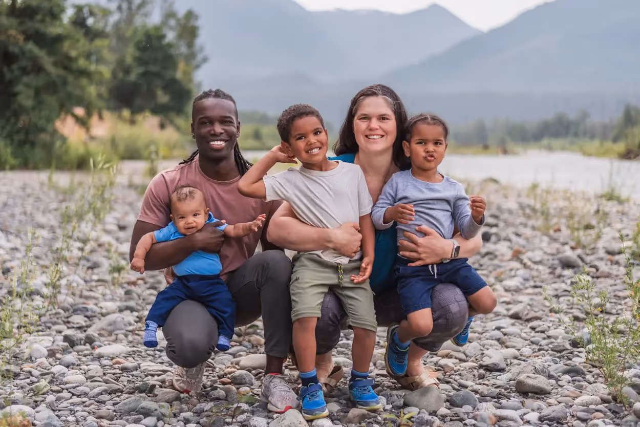 Smiling family of five posing on a rocky riverbed with mountains in the background.