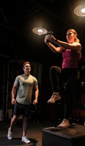Woman doing step-up exercise with kettlebell while a man watches in a gym setting under overhead lights.