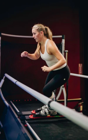 Young woman in a white tank top and black leggings skiing on an indoor ski simulator with handrails.