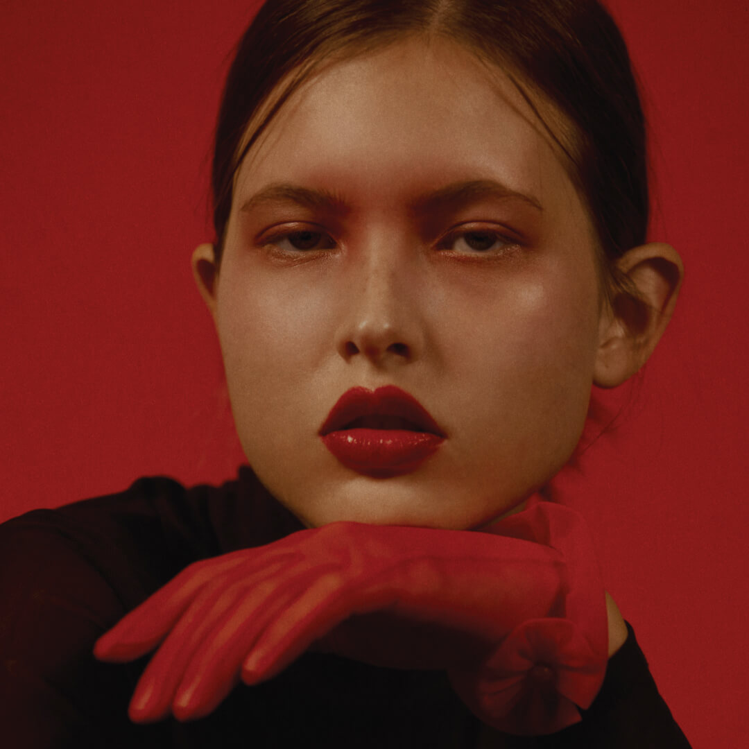 brunette model with dark red lips staring into camera with a bored expression on her face and her chin resting on her red gloved hand the background is red color