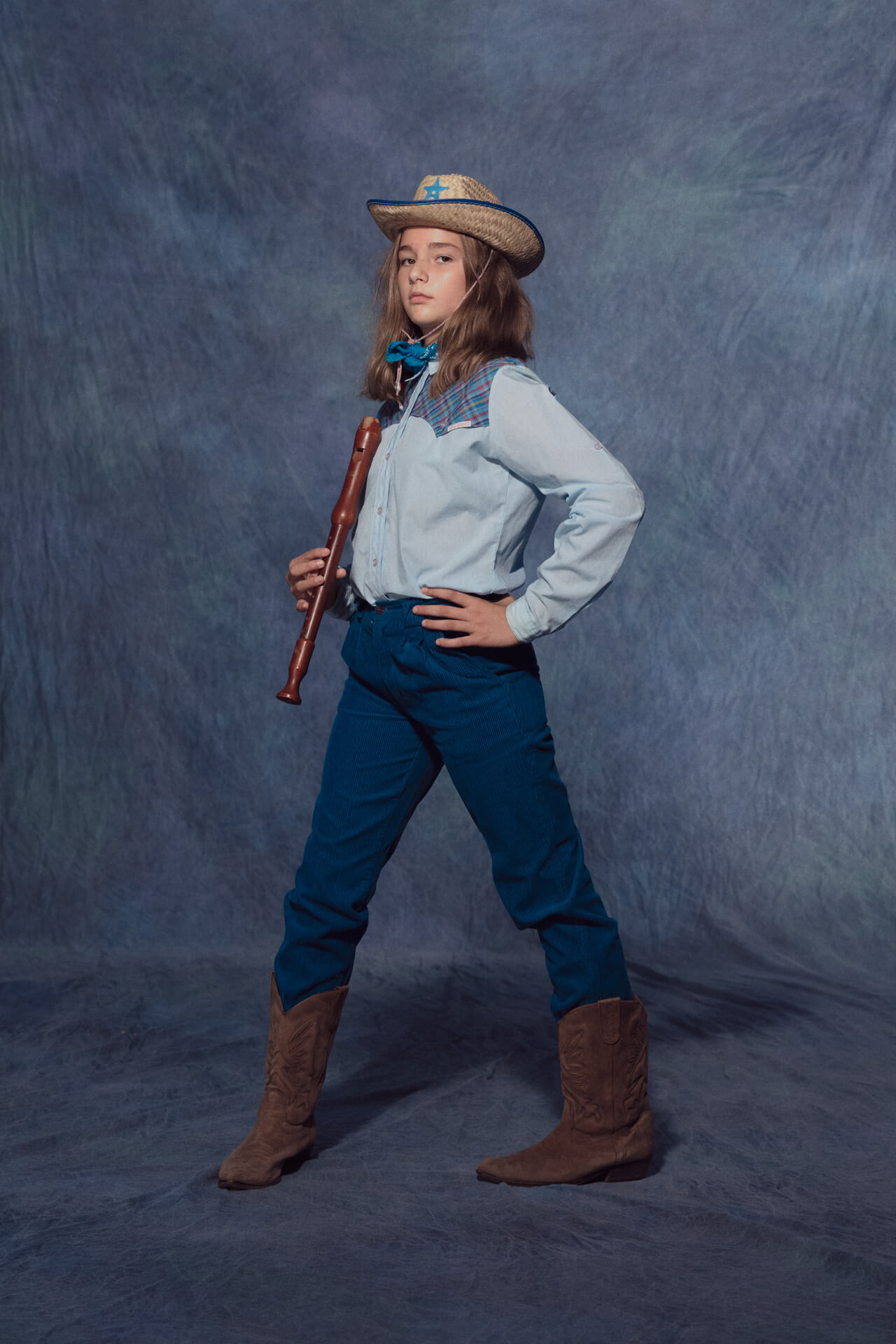 Young girl from the americana series holding a recorder instrument and wearing a cowboy hat and a hand on her hip staring at the camera