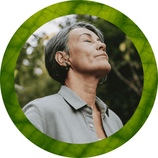 A person with short gray hair stands outdoors with eyes closed and face tilted upward, wearing a light gray collared shirt. The portrait is framed by a thick green circular border.