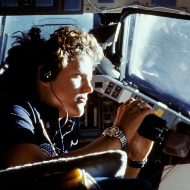 Kathryn Sullivan wearing a headset sits inside a spacecraft cockpit, holding a small camera or instrument near the window.