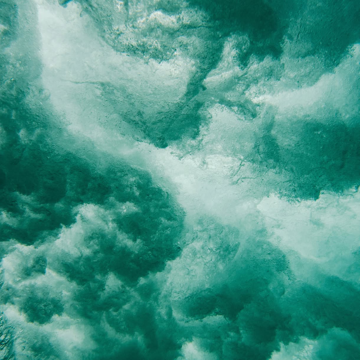 A turbulent underwater view of breaking waves fills the frame with swirling turquoise water, bubbles, and white foam.