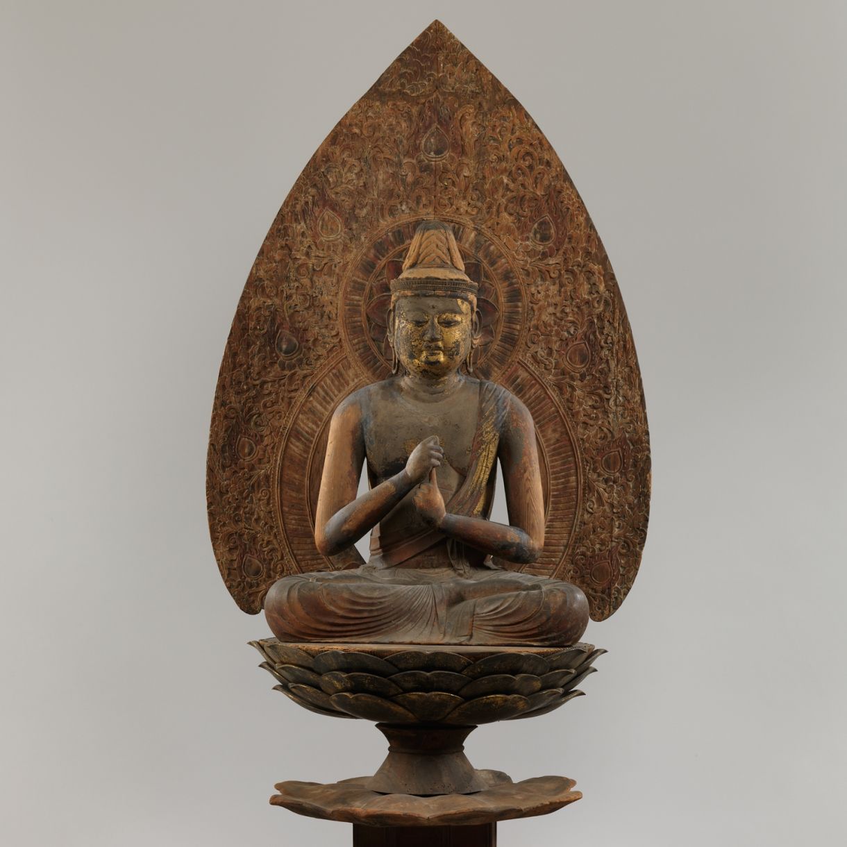 A Japanese Buddhist sculpture depicting a seated Buddha on a lotus pedestal, framed by a tall, intricately carved mandorla.